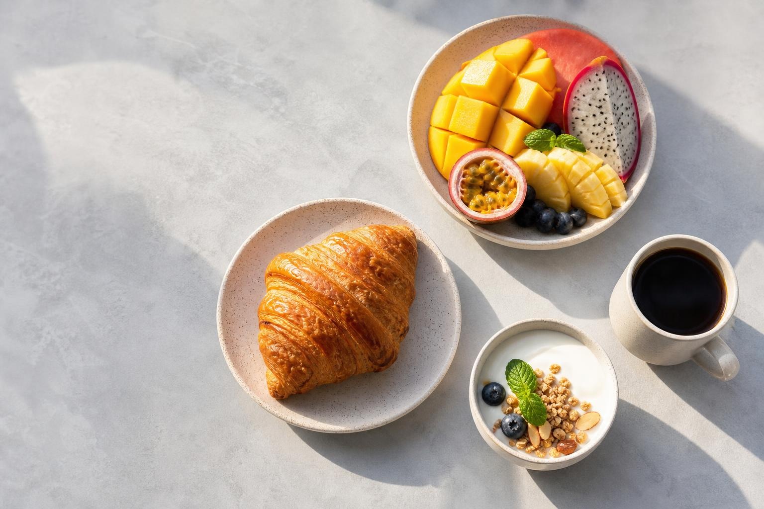 Illustrative overhead brunch spread with tropical fruit, croissant, yogurt and coffee.
