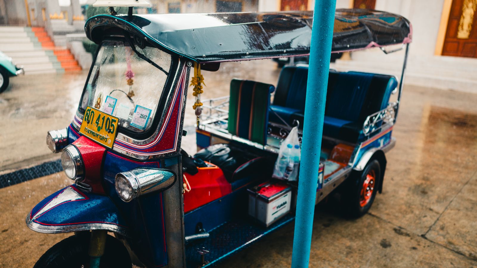 Illustrative close view of a colorful tuk-tuk parked on a wet Bangkok street.