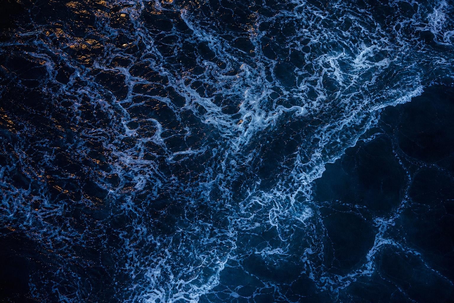 Illustrative overhead view of dark blue ocean waves with white foam at night.
