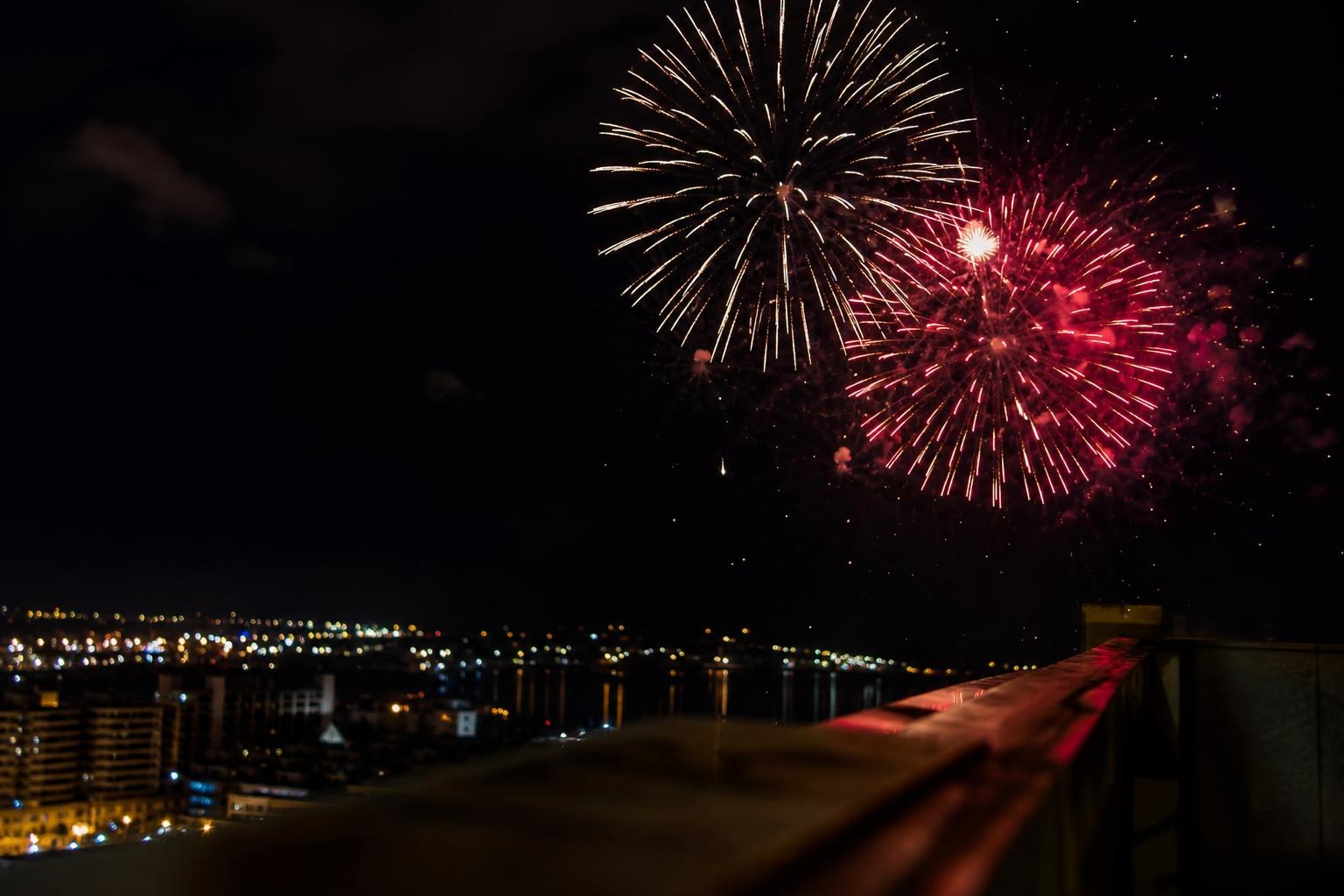 Illustrative gold and red fireworks above a distant waterfront city, seen beside an unbranded rooftop railing.